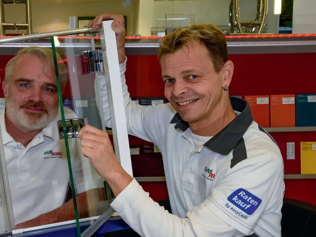 Die Gründer Dirk Artz und Michael Schönfelder in weißen Glasteufel-Poloshirts an einer Ganzglastür mit Stangengriff, dahinter ein Regal mit Farbmustern